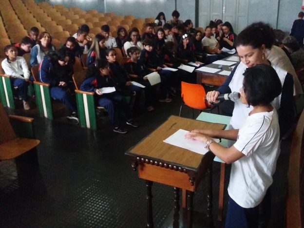 Descrição da imagem: foto de mulher segurando microfone na altura da boca de uma moça que tateia papel em branco. À frente delas há um grupo de pessoas sentadas em cadeiras universitárias.